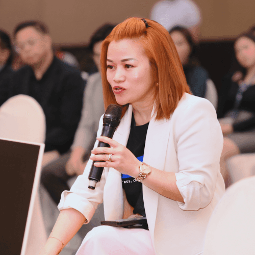 Person in a white blazer holding a microphone, sitting among an audience at an indoor event.